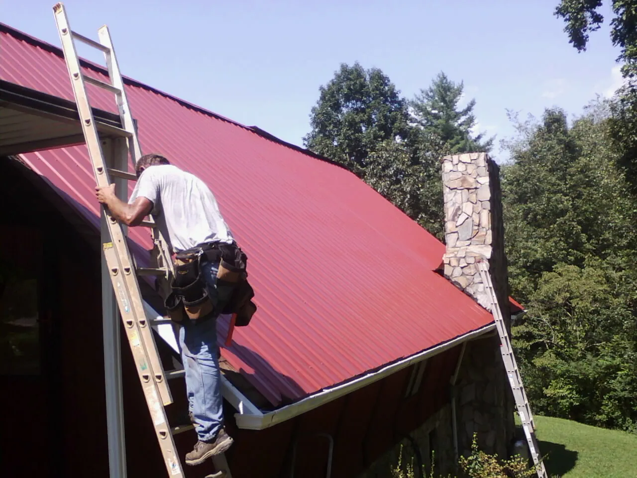 Roofer on a ladder beside a red metal roof and stone chimney — Soffit & Fascia Repair by Roofing Brothers in Millersburg